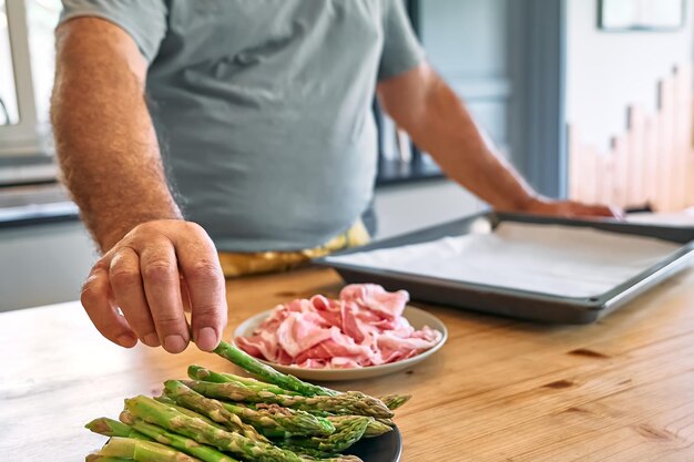 Foto man bereidt jonge groene aspergespruiten voor die in spek zijn gewikkeld op een houten tafel in de keuken