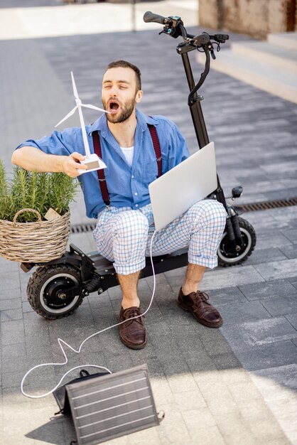 Man blaast op speelgoedwindgenerator terwijl hij buiten met laptop en groene plant op elektrische scooter zit