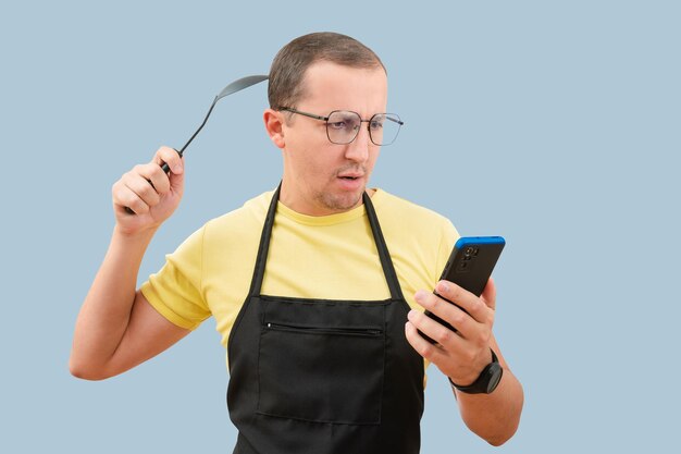 Man in black apron holding spoon using smartphone in hand isolated on blue background