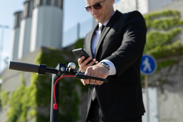 Man in a black business suit stands next to an electric scooter and holds a phone