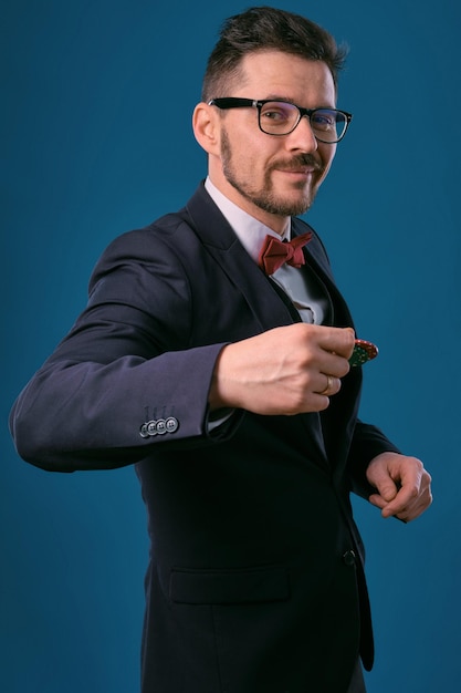 Man in black classic suit red bowtie glases is showing two colored chips posing on blue studio backg...