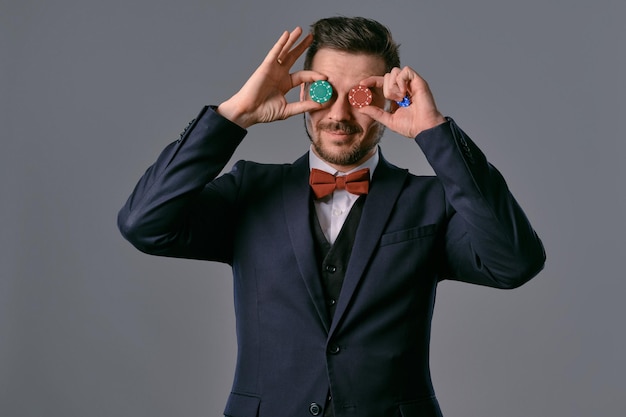 Man in black classic suit and red bowtie showing some colored chips posing against gray studio backg...