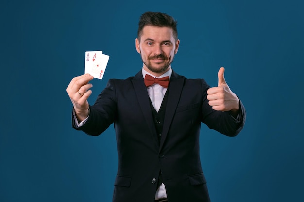Man in black classic suit and red bowtie showing two playing cards while posing against blue studio ...