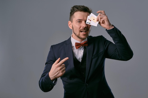 Man in black classic suit and red bowtie showing two playing cards while posing against gray studio ...