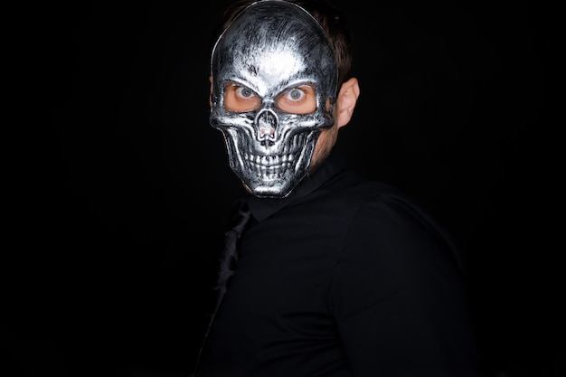 A man in black clothes stands on a black background wearing a skeleton mask