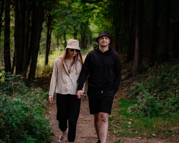 a man in black clothes and a woman in a beige shirt and hat are walking holding hands in the forest