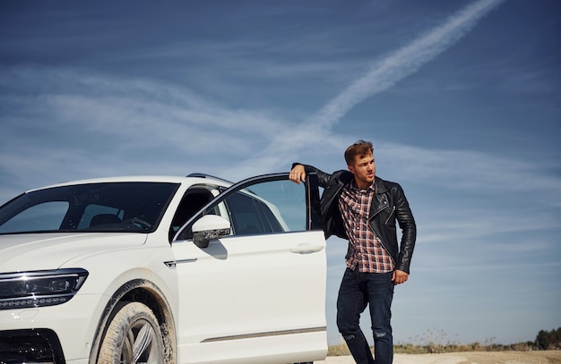 Man in black leather jacket stands near his parked white car outdoors against blue sky.