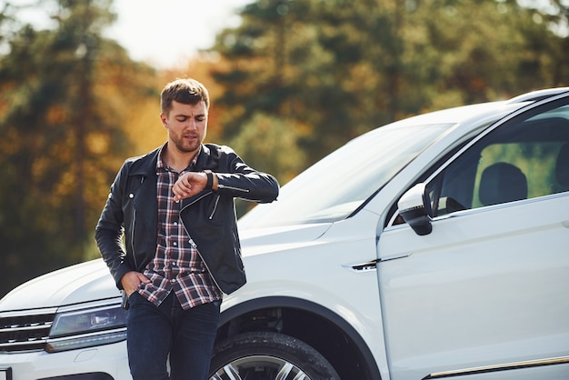 Man in black leather jacket stands near his parked white car outdoors and waits for help.