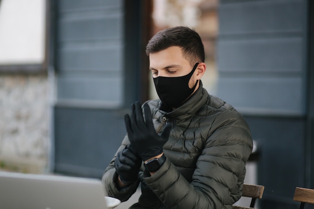 昼食をとるために黒い手袋をはめた黒い保護マスクの男。検疫covid-19の間にカフェで屋外に座っている保護マスクの男。