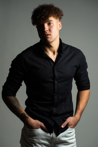 A man in a black shirt and jeans stands in front of a grey background.