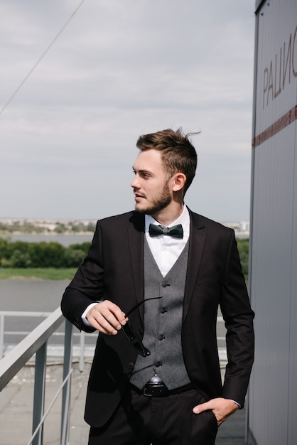 a man in a black suit and a black bowtie poses in the open air.