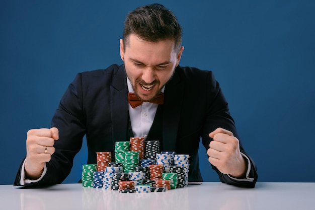 Man in black suit is sitting at white table with colored stacks\
of chips on it posing on blue studio...