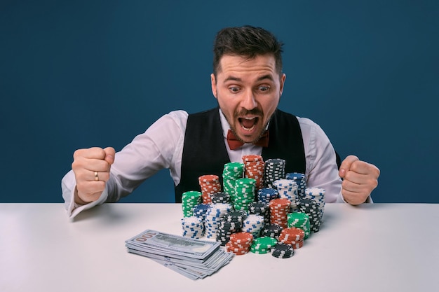 Man in black vest and shirt sitting at white table with stacks of chips and cash on it posing on blu...