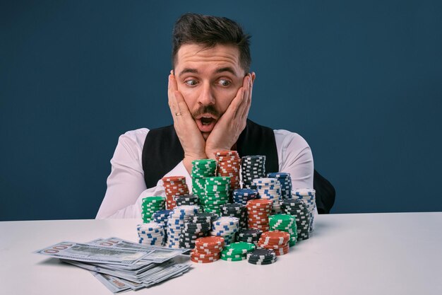 Man in black vest and shirt sitting at white table with stacks of chips and cash on it posing on blu...