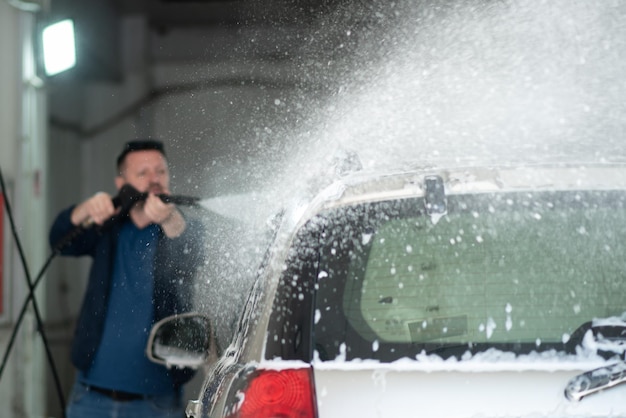 A man in a blue jacket and sunglasses washes his car at a selfservice car wash