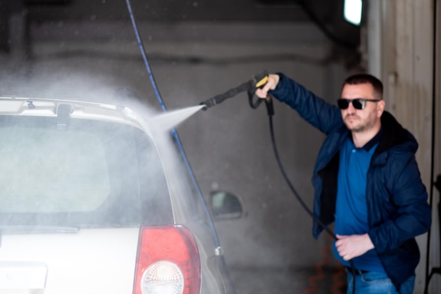 A man in a blue jacket and sunglasses washes his car at a selfservice car wash