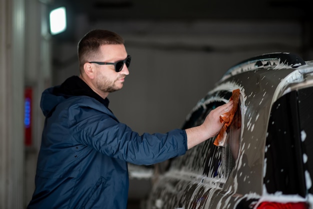 A man in a blue jacket and sunglasses washes his car at a selfservice car wash