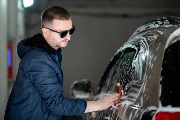 A man in a blue jacket and sunglasses washes his car at a selfservice car wash