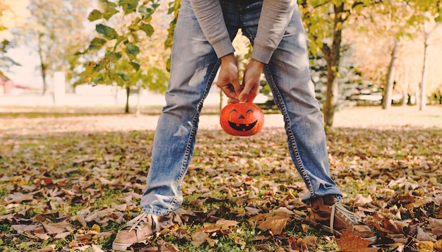 A man in blue jeans holds a pumpkin between his legs in a Park among yellow leaves.