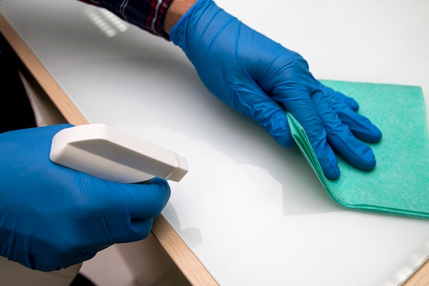 A man in blue protective gloves disinfects the surfaces using a rag and spray disinfectant