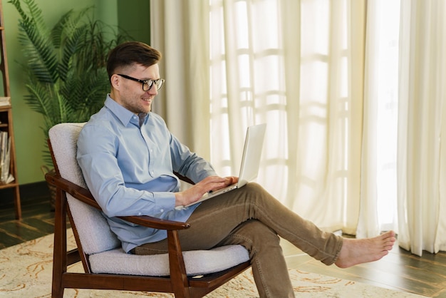 Man in blue shirt and brown pants comfortably working on a laptop barefoot in a cozy room