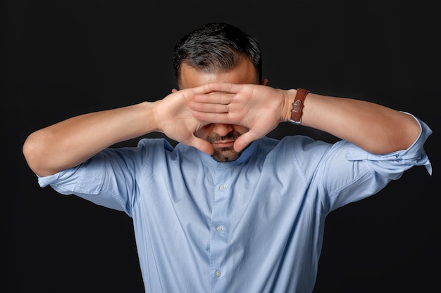 Man in blue shirt covers his face with his hands on black background.