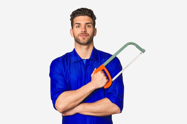 A man in a blue shirt is holding a saw with his arms crossed.