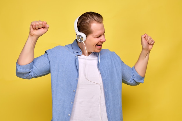 A man in a blue shirt on a yellow background dressed in white headphones and enjoys dancing music.