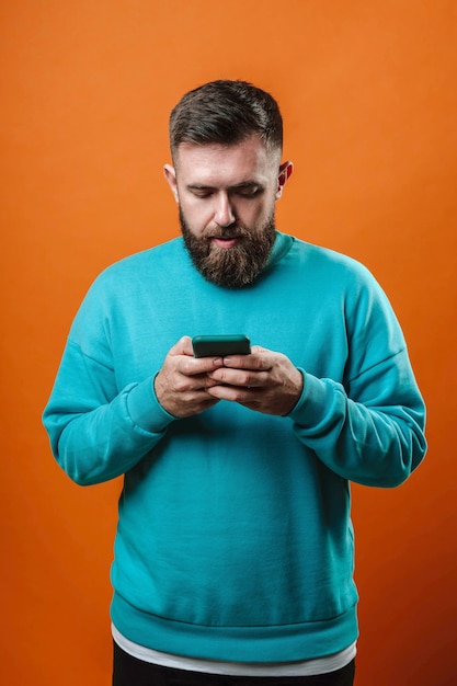 Человек в синем свитере с мобильным телефоном в руках на оранжевом фоне студии