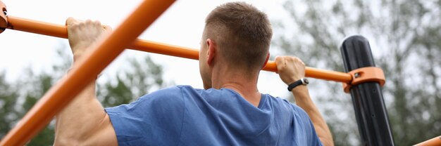 Man in blue t-shirt pull himself up on horizontal bar in park. Inflated athlete do exercise on sports equipment.