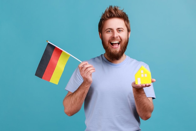 Man in blue Tshirt holding toy paper house and flag of germany in hand moving into another country