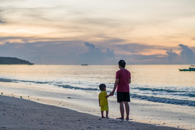 A man and a boy stand on the beach, holding hands, looking out to the ocean.