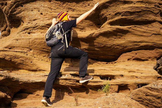 Photo man brave red-haired beard climbing bouldering stone rock tourist climbs up with backpack in a black t-shirt and a funny hat made of wool of yak from nepal