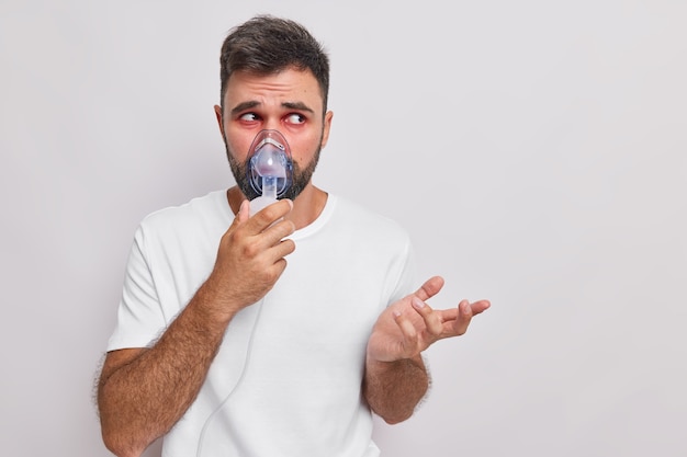  man breathees through nebulizer mask makes therapeutic procedures treats respiratory disease or allergy suffers from asthma shrugs shoulders and looks clueless isolated on white studio wall