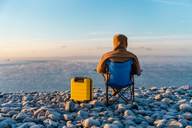 Man in brown jacket with yellow suitcase relaxing alone and sitting on beach chairs on seaside