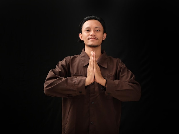 A man in a brown shirt is standing with his hands together in prayer.