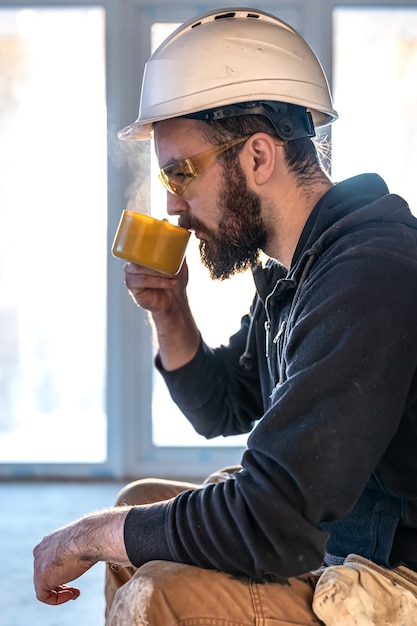 A man builder in a helmet and goggles drinks a hot drink.