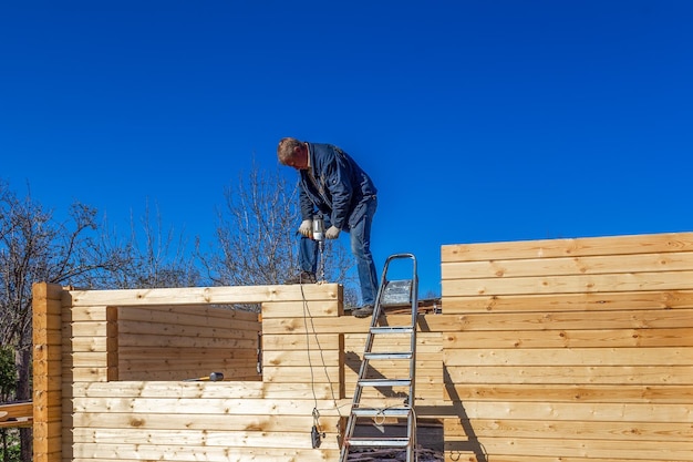 A man builds a house made of profiled timber in the countryside against the blue sky