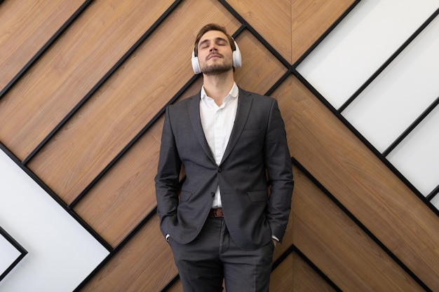 A man in a business suit on the background of an office wall in headphones listens to music with