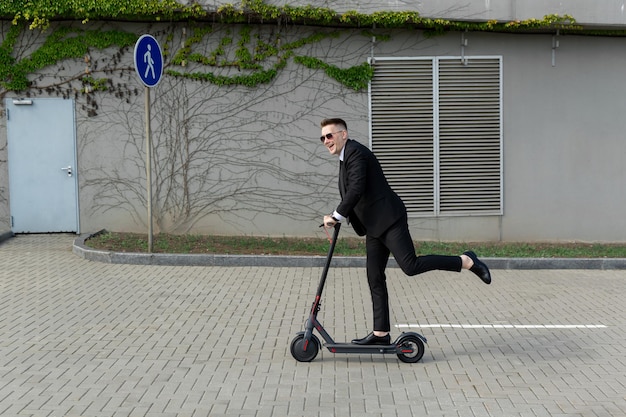 Man in a business suit and sunglasses rides an electric scooter on one and laughs