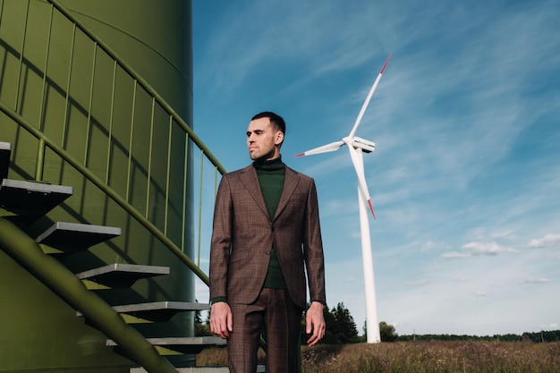 A man in a business suit with a green Golf shirt stands next to a windmill against the background of the field and the blue sky.Businessman near the windmills.Modern concept of the future.