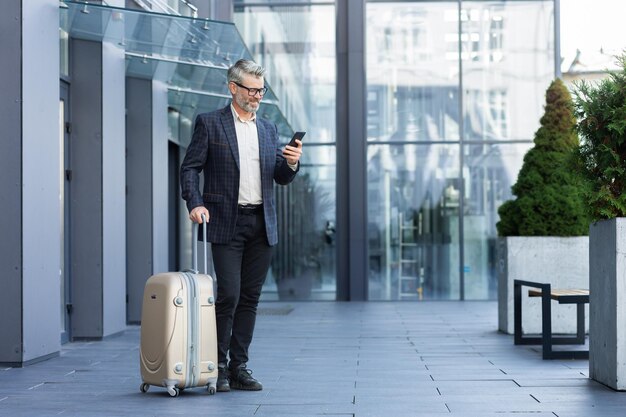 A man in a business suit with a large travel suitcase and a phone is typing a message and booking a