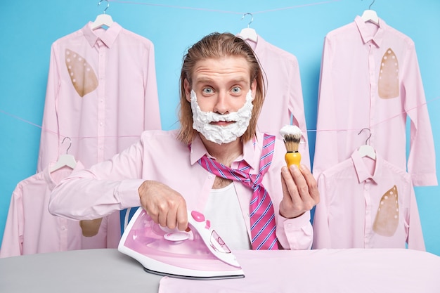 Man busy shaving and ironing washed folded clothes after\
laundry does everyday domestic activities holds brush uses electric\
steam iron being in hurry for work