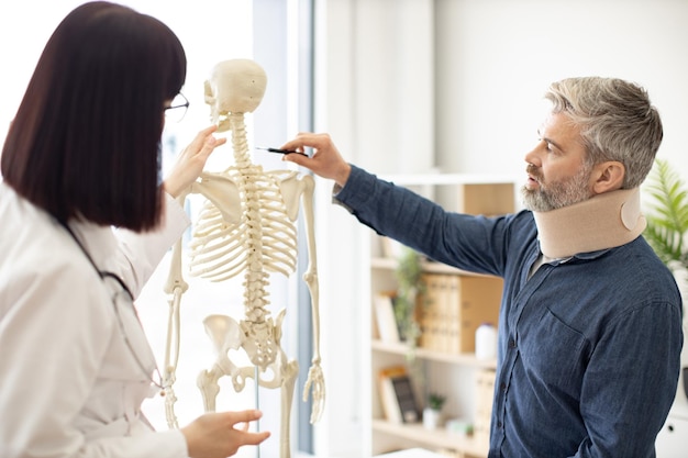 Man in C collar asking physician about spine using skeleton