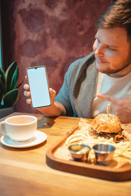 Man in cafe holding phone with white screen eating burger\
drinking tea