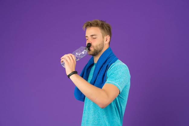 Man on calm face drinking water after training in gym violet background guy with bristle wears fitness tracker sporty lifestyle concept man with towel on neck renews water balance after training