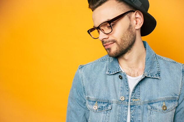 Un uomo di umore calmo con un leggero sorriso indossa una giacca di jeans, una t-shirt bianca, occhiali semplici e un cappello nero sui suoi lucenti capelli castano scuro