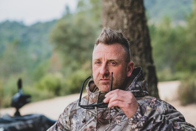 Foto un uomo con una giacca mimetica tiene i suoi occhiali da sole davanti a un albero.