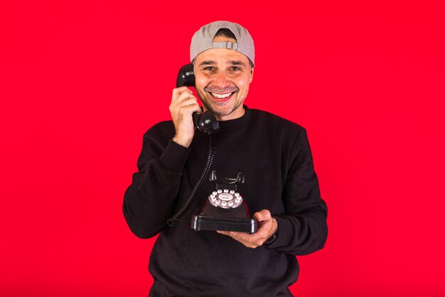 Man in a cap dressed in black, answers a call with a retro telephone from the 60s and smiles, on a red background, with copy space. Concept of contact, social relations and phone calls