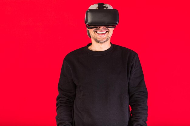 Man in cap dressed in black wearing virtual reality glasses, smiling, on red background. technology, vr, computing and hobbies concept